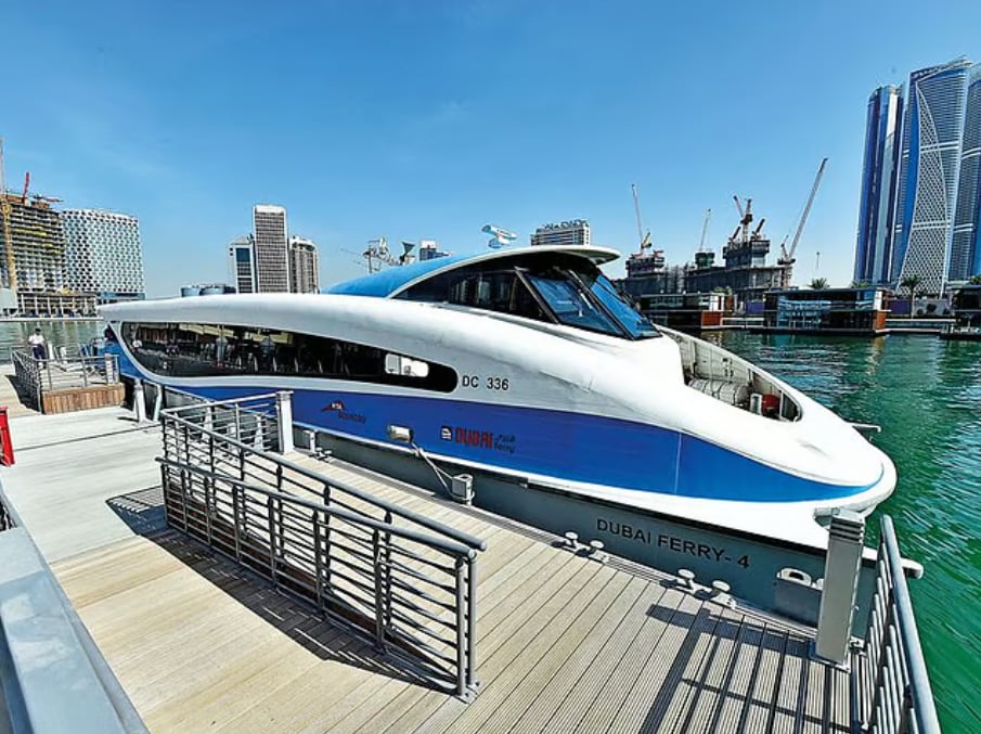 A blue and white Dubai Ferry moored at a wooden jetty with towers behind