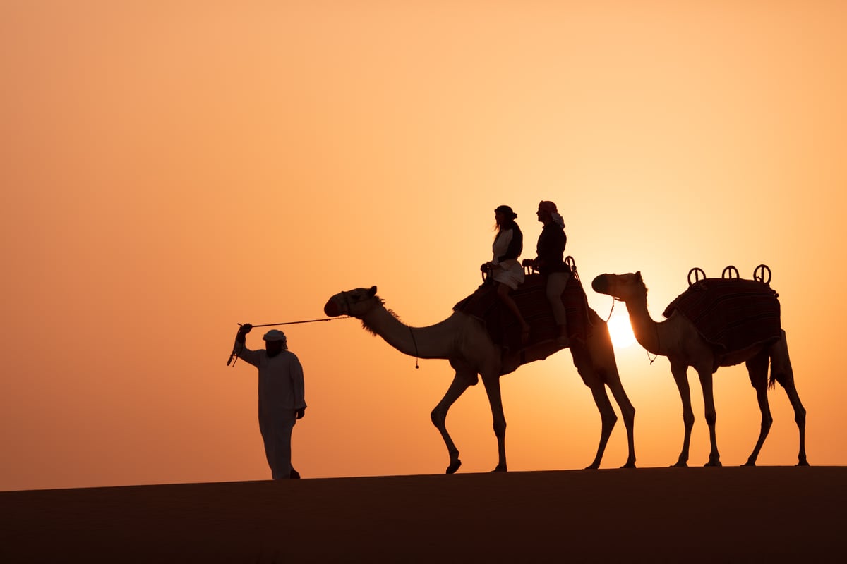 A guide leading two riders on camels, silhouetted against an orange sky