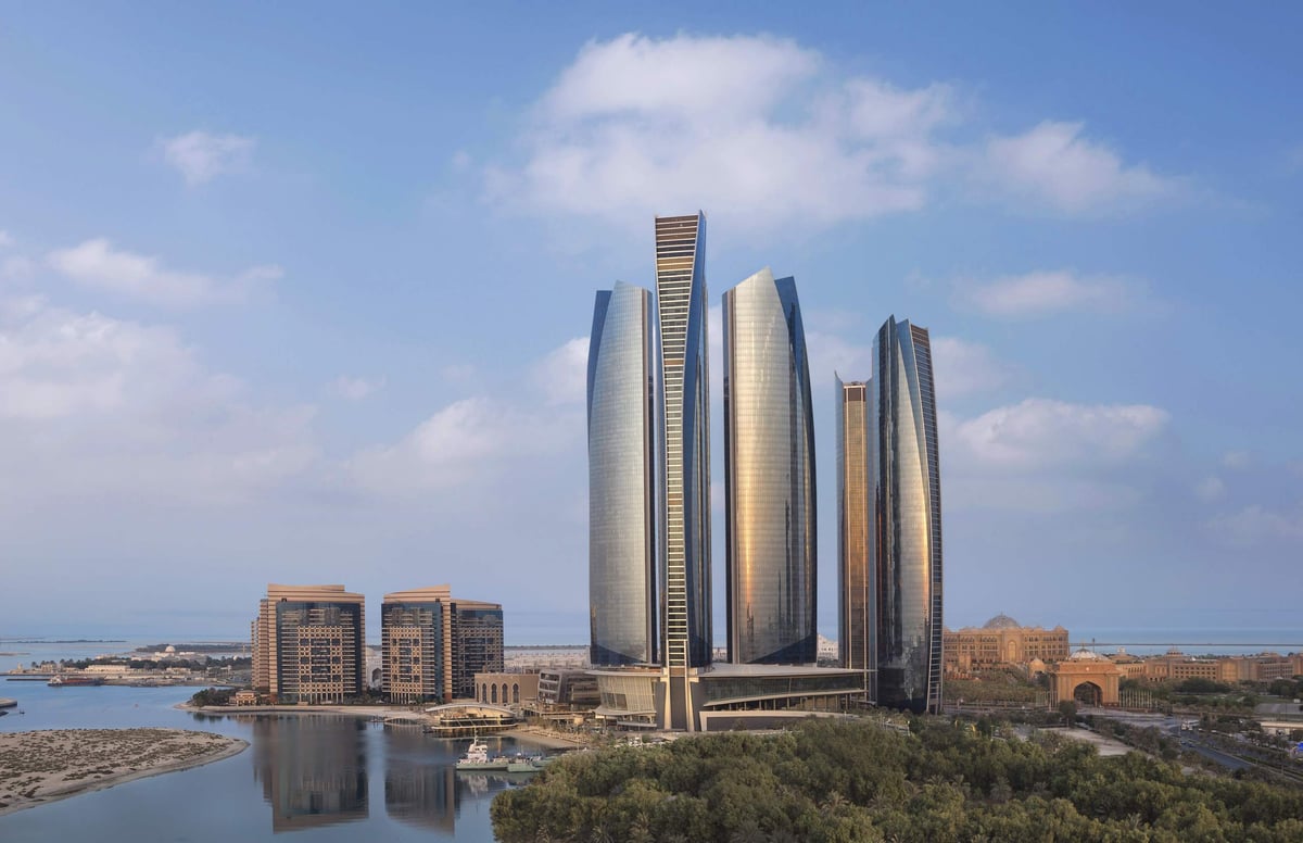 The five curved glass towers of Conrad Abu Dhabi Etihad Towers rising behind a calm inlet and low waterfront buildings under a blue sky.