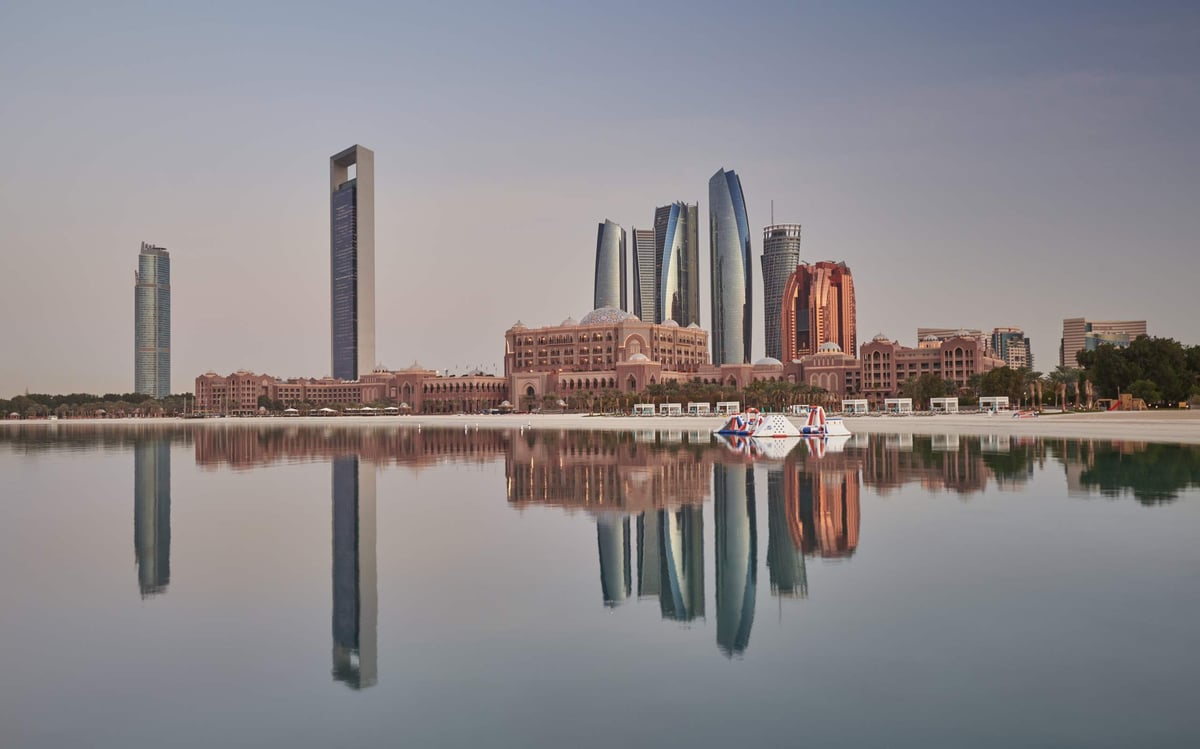 The full length of Emirates Palace and the Abu Dhabi skyline mirrored in calm water on a clear day.