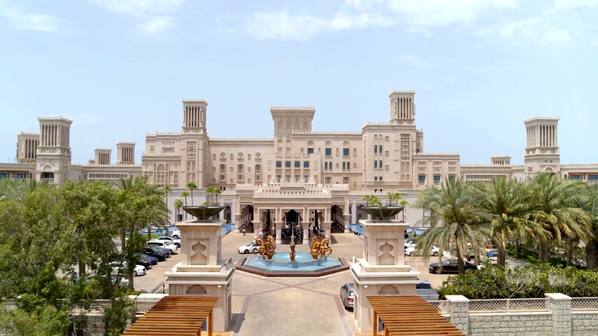 Drone footage over Jumeirah Al Qasr: the entrance bridge and forecourt, palm gardens and pools, canals, and a couple walking the beach pier.