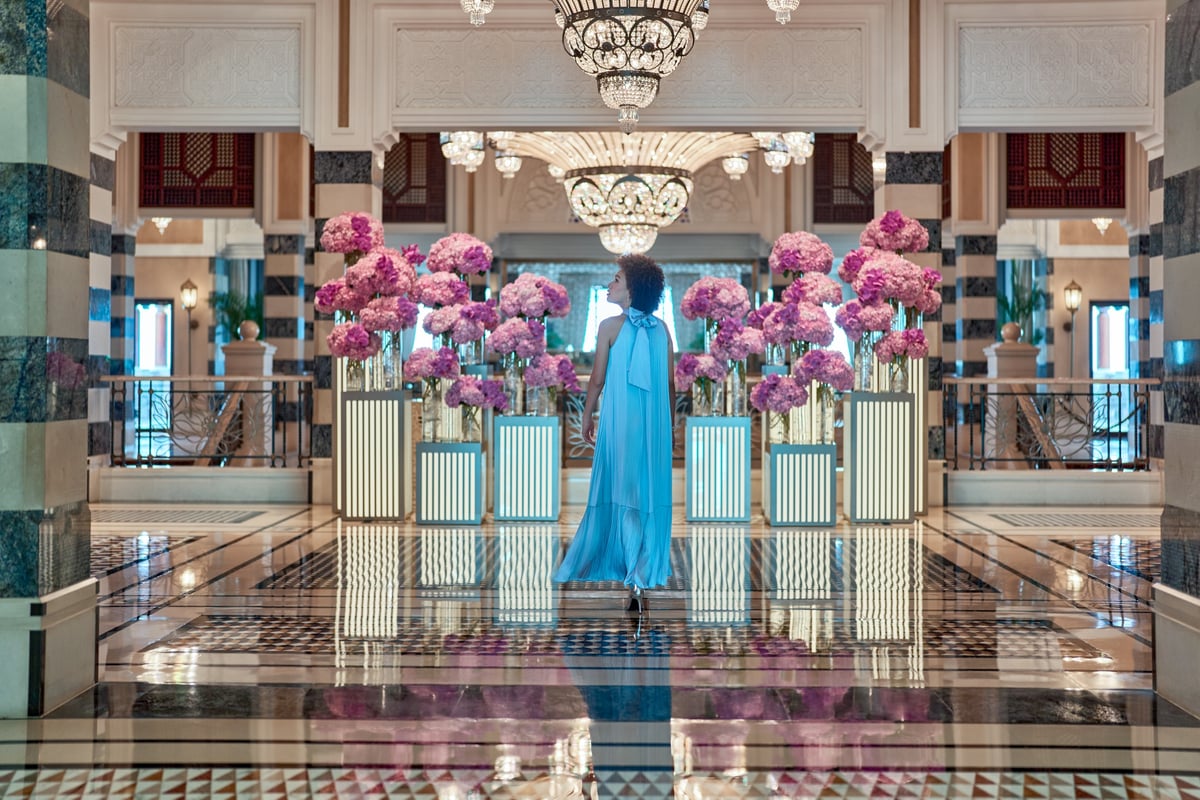 A woman in a blue gown stands among pink flower arrangements under a chandelier in the Jumeirah Al Qasr lobby.