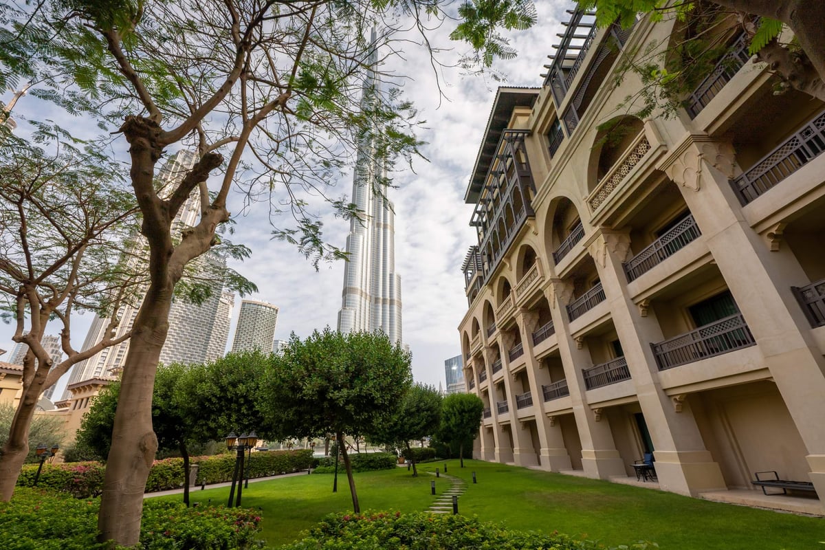 A lawn with trees beside Palace Downtown's arched balconied wing, with Burj Khalifa towering in the background.
