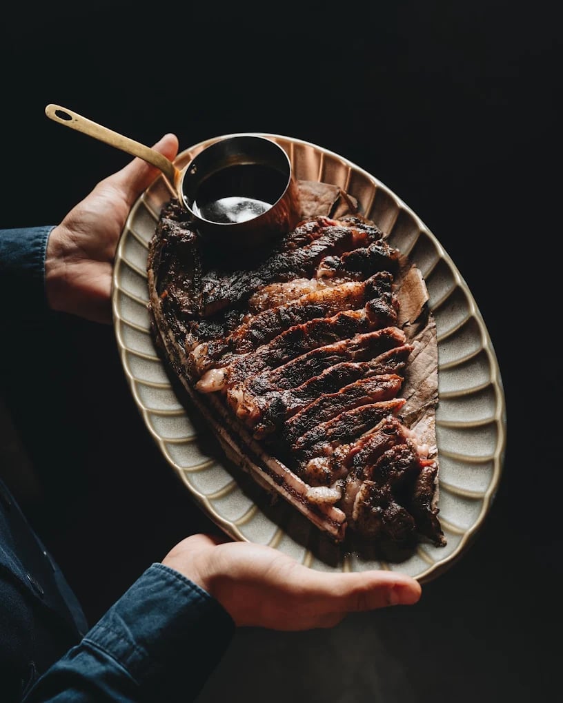 A sliced chargrilled steak on a ridged oval platter held out with a sauce ladle