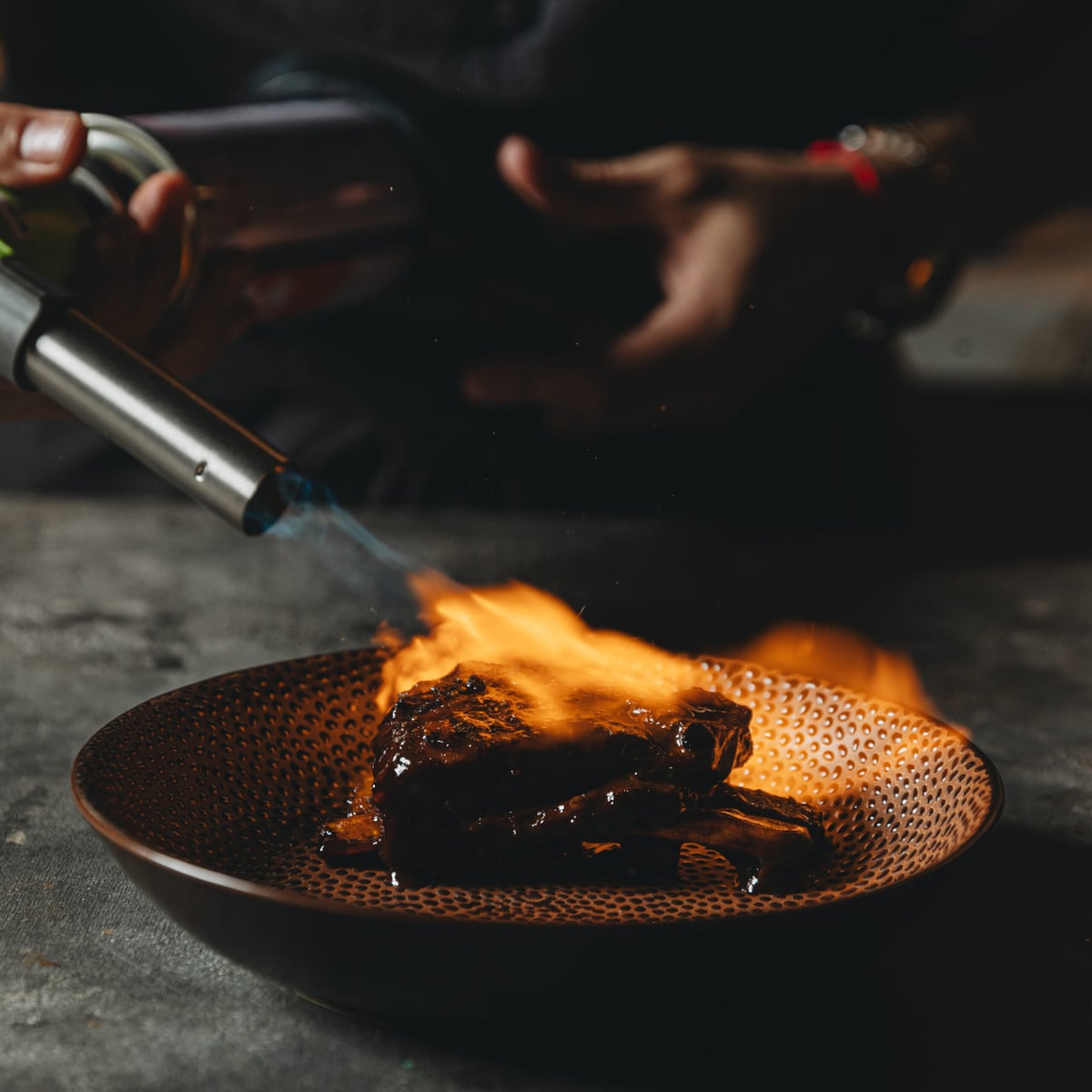 A chef torching a glazed cut of meat in a hammered copper bowl, flames rising from the plate