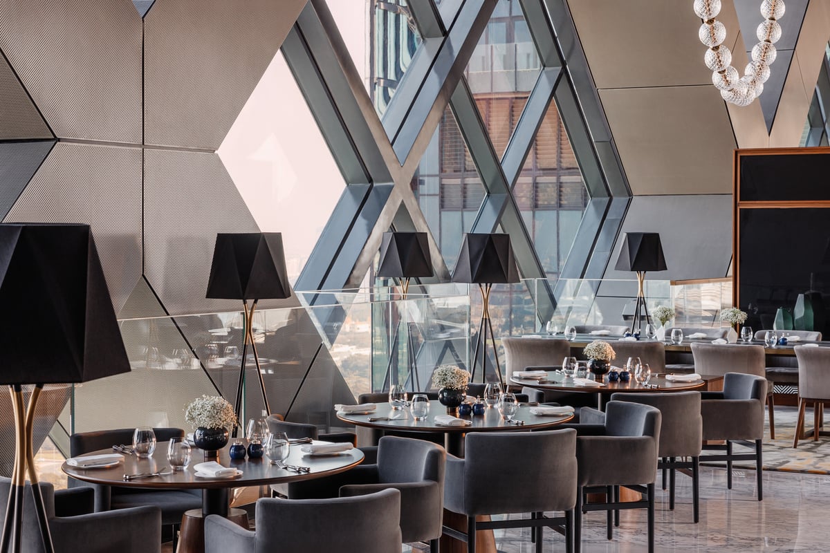 Tables laid for dinner against the diagonal steel and glass windows, black shaded lamps along the wall