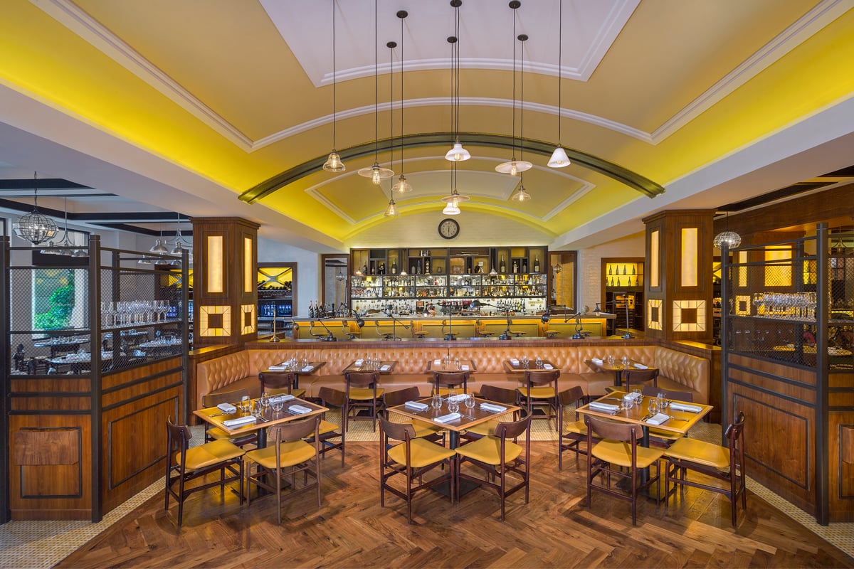 Wooden tables and yellow-upholstered chairs beneath a vaulted ceiling washed in yellow light, the bar at the far end