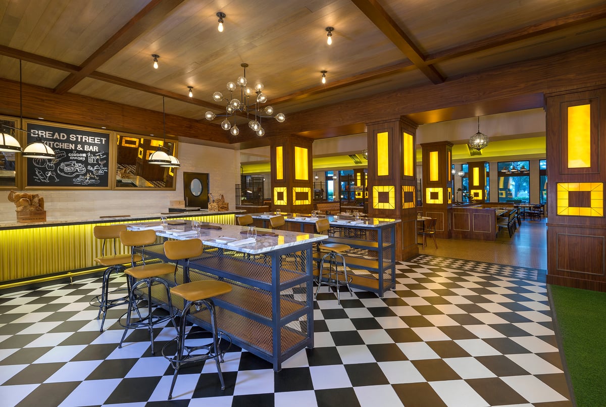 High tables with stools on a black-and-white checkerboard floor beneath a beamed ceiling and hanging bulbs