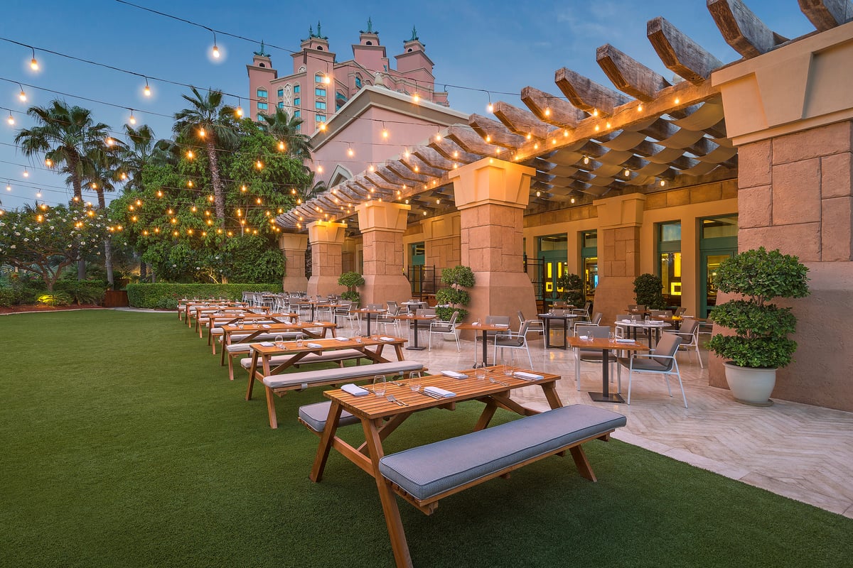 Long wooden picnic tables on a lawn under strings of lights at dusk, Atlantis The Palm's towers behind the palms
