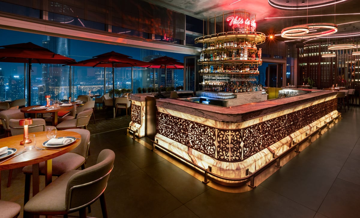 The illuminated bar glowing gold at night beneath tiered shelves of bottles, terrace parasols beyond