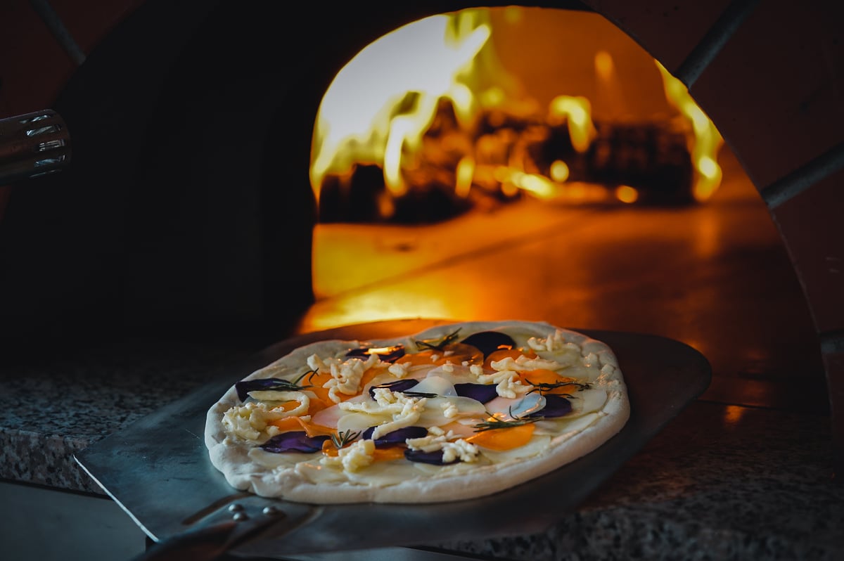 A pizza on a peel in front of the flames of the wood-fired oven