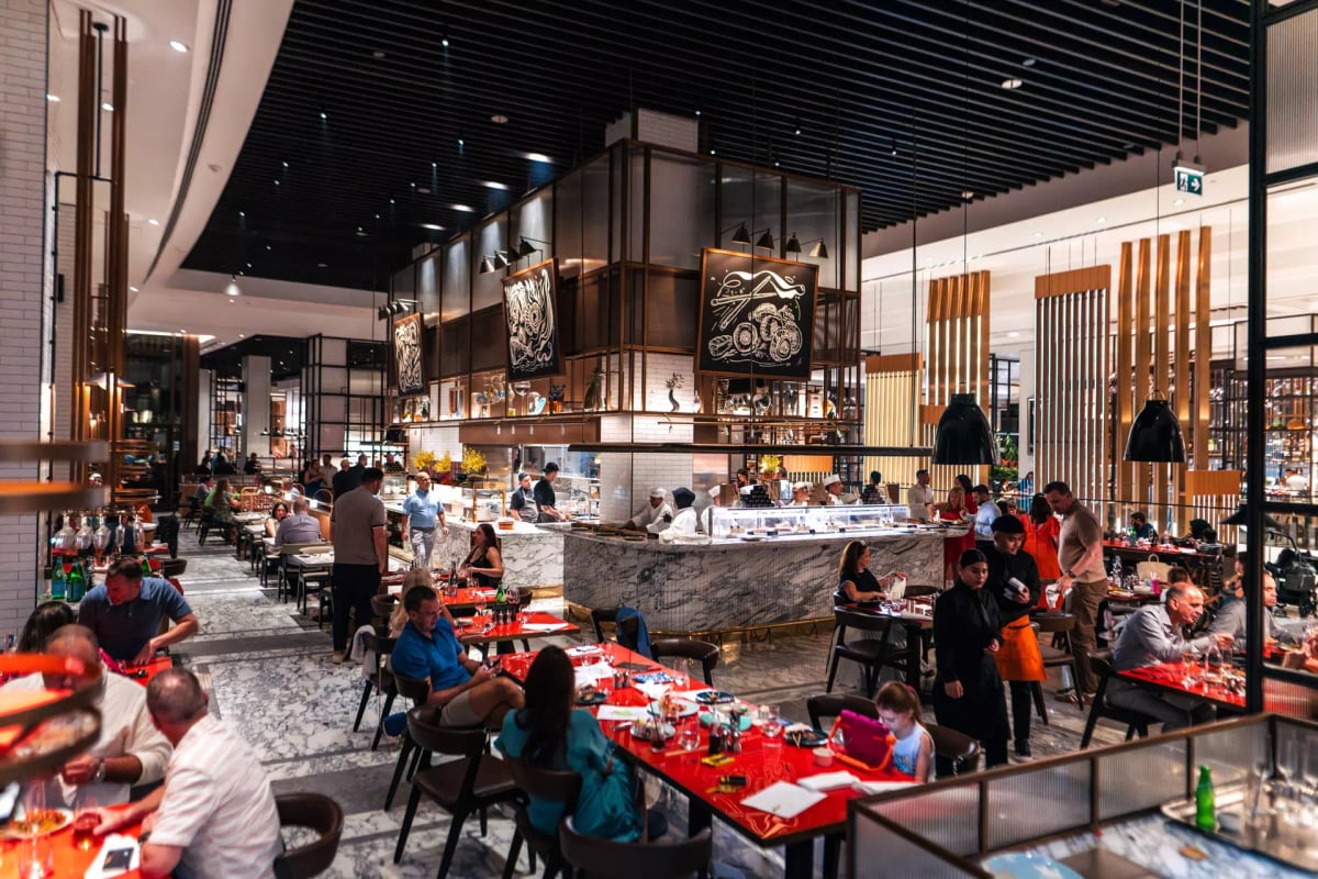 Guests dining at red tables across a busy food hall of marble counters under a dark slatted ceiling