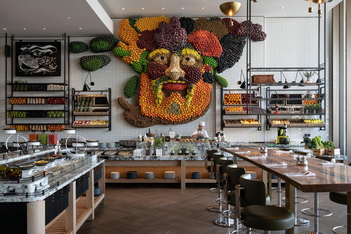 A giant face made of fruit and vegetables on a white tiled wall above a salad counter and long high table