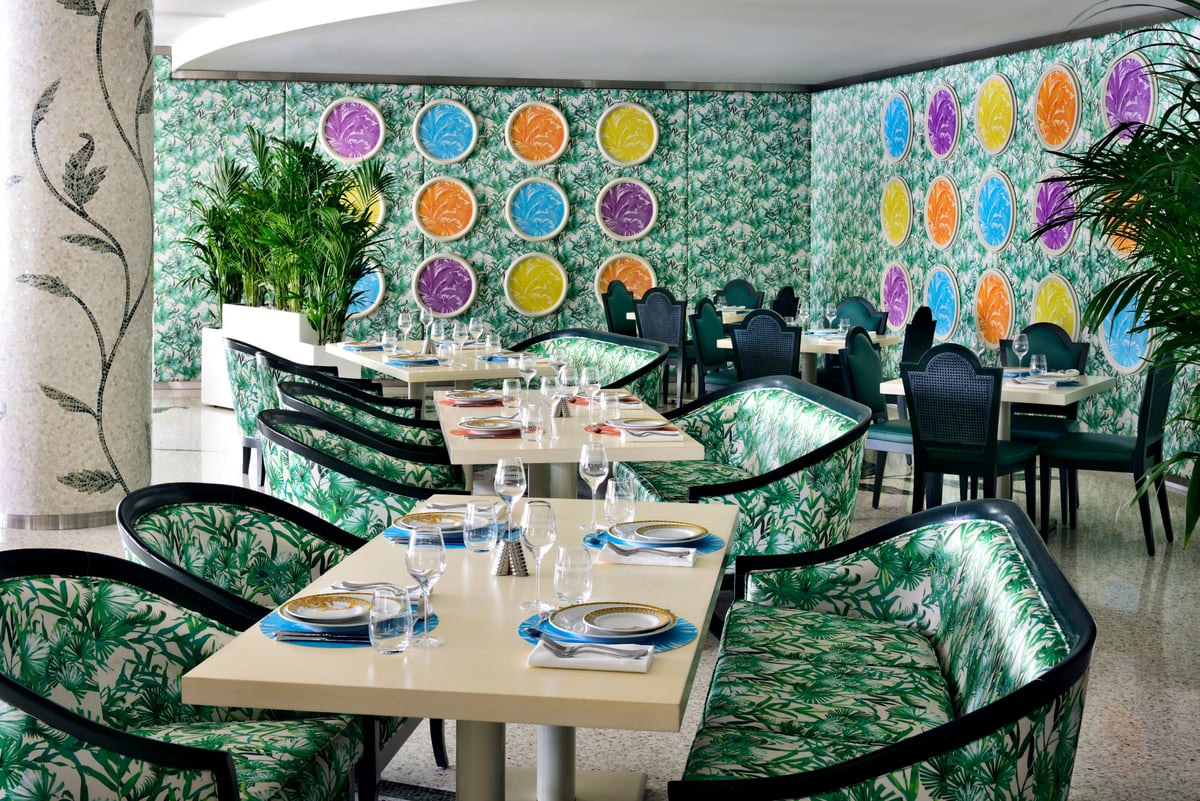 Tables with palm-print chairs in front of a wall of colorful decorative plates on green botanical tiles
