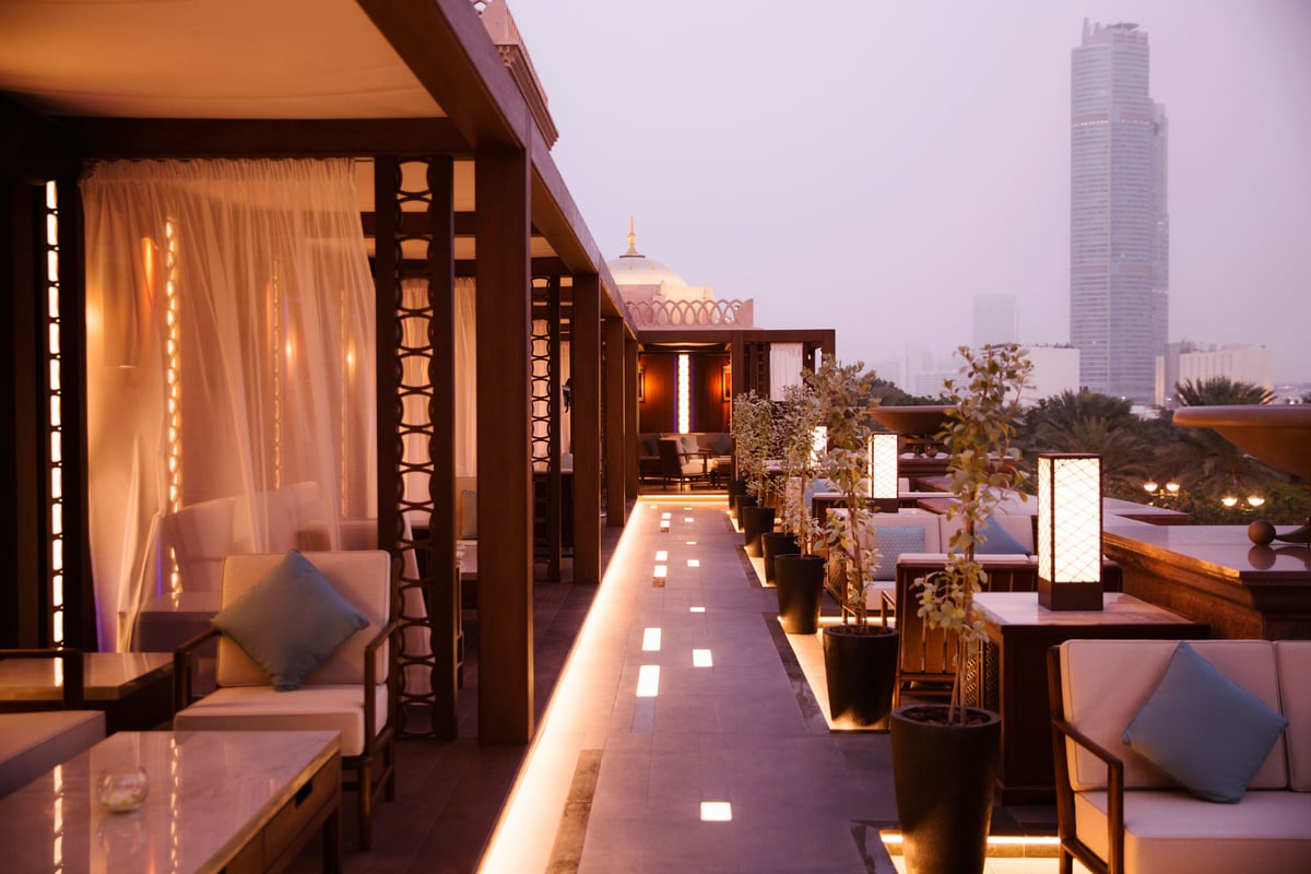 A terrace walkway lit along the floor between curtained cabanas and planters, a tower in the distance