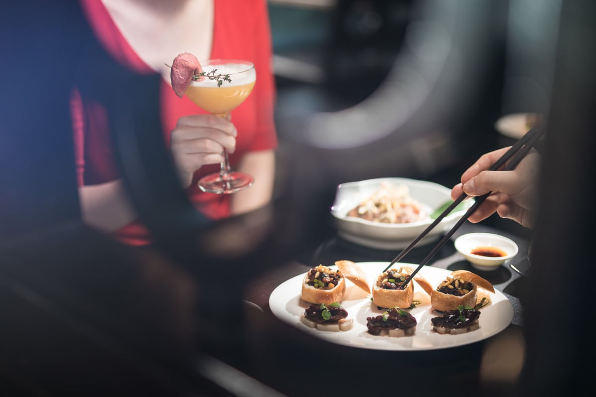 A guest lifting a dim sum parcel with chopsticks beside a cocktail garnished with a dried flower