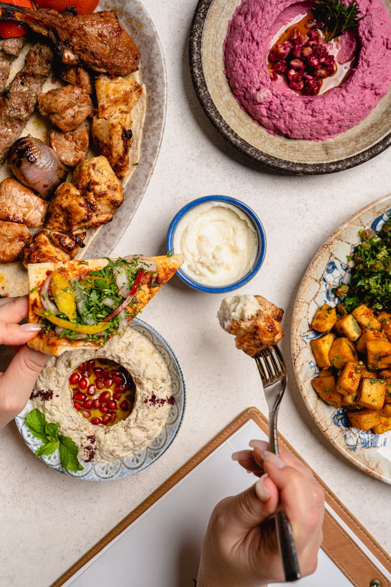 Hands reaching across a table of beetroot hummus, labneh, grilled meats and flatbread