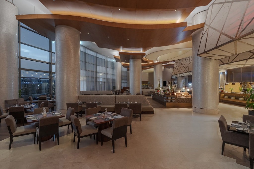 Tables and dark chairs among white columns, floor-to-ceiling windows along one side