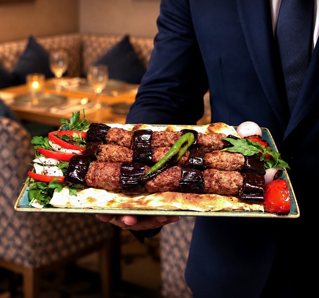 A waiter carrying a platter of lamb kebabs and grilled aubergine on flatbread with salad