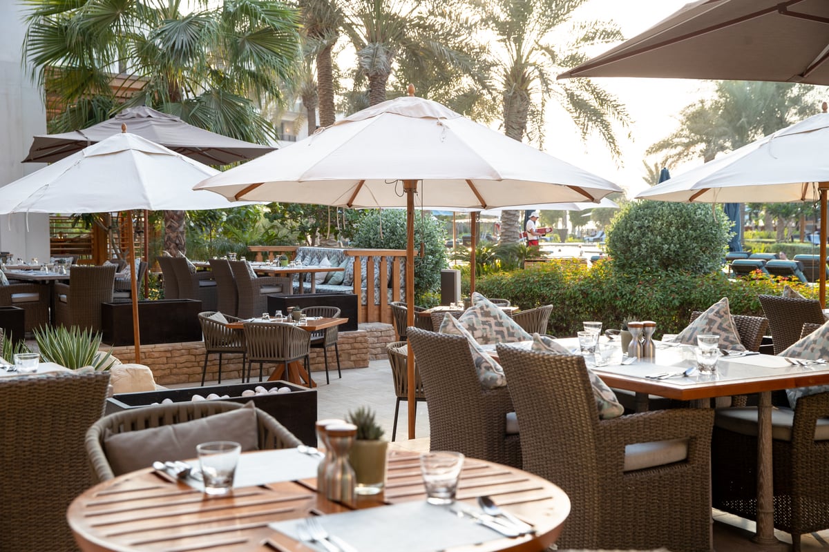 Wicker armchairs and round timber tables under white parasols, palms and gardens behind