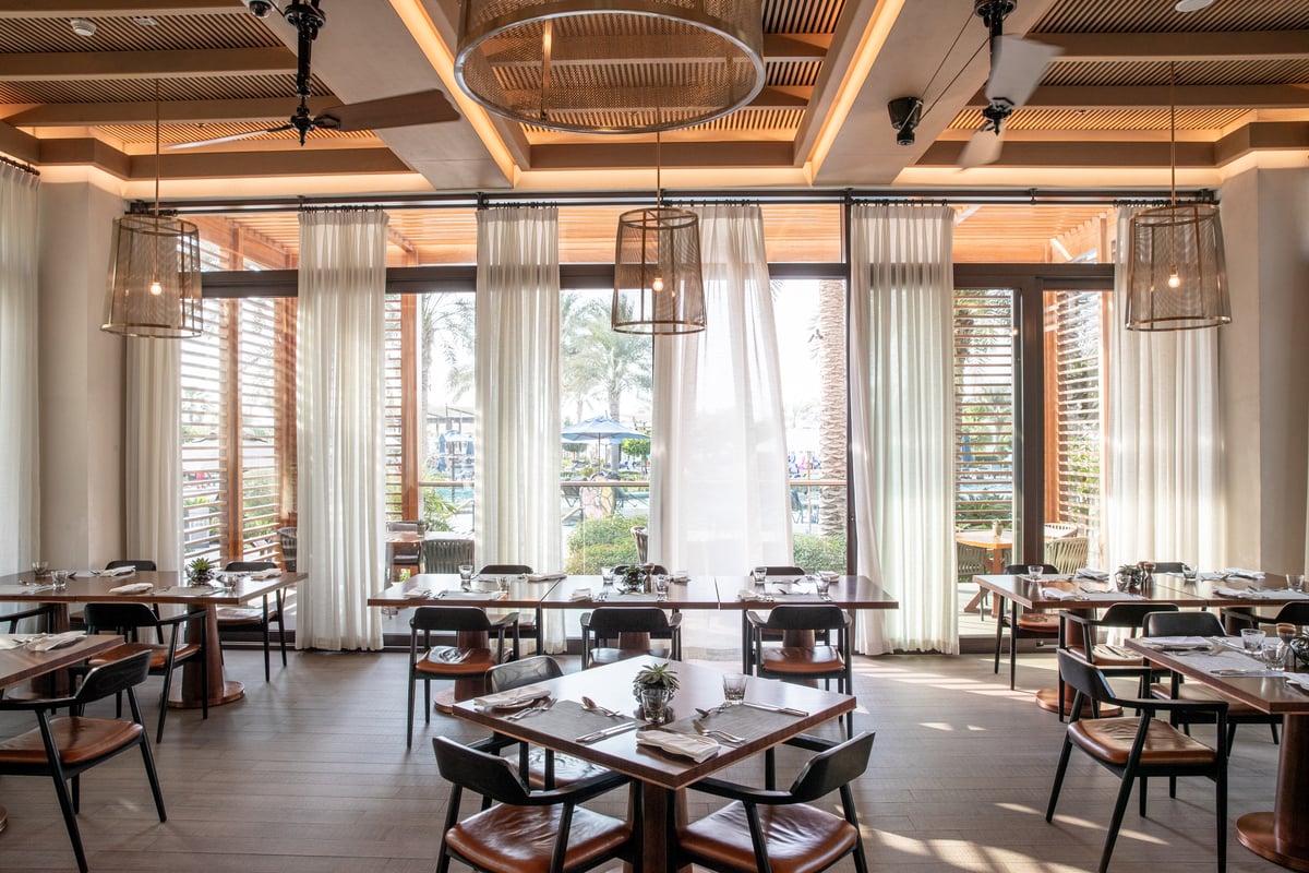 Tables and leather-seated chairs beneath a slatted timber ceiling, the garden glimpsed through open shutters