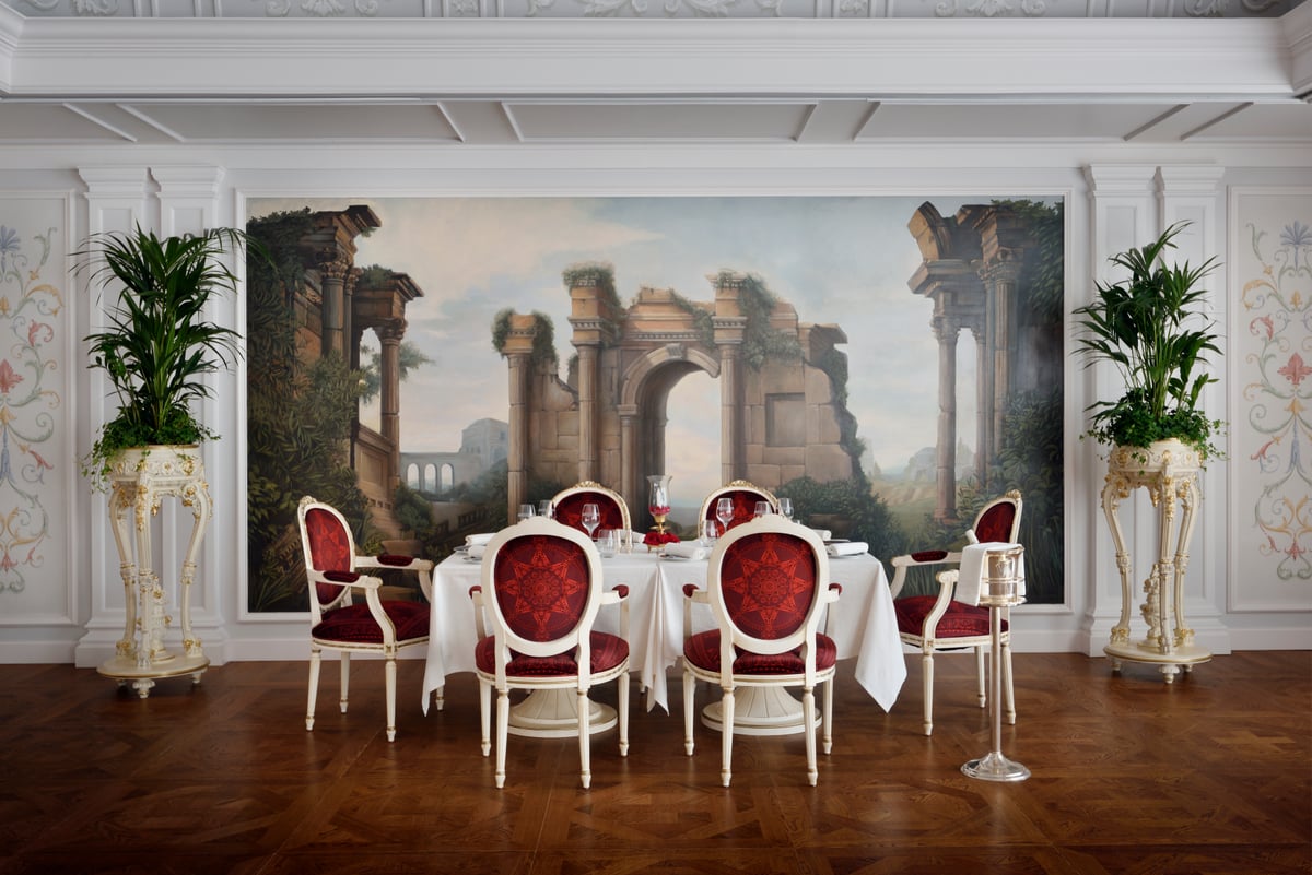 Red-upholstered gilt chairs at a laid table before a large mural of classical ruins