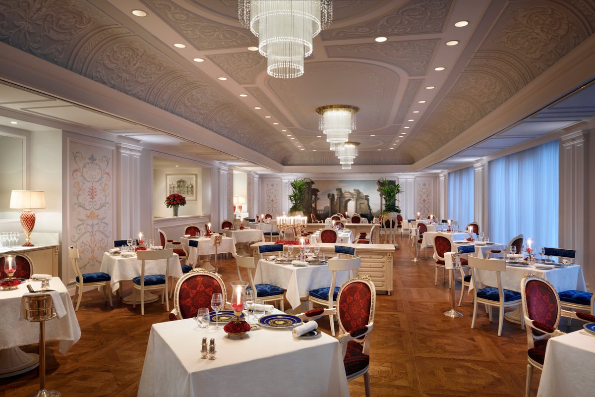 White-clothed tables and red-and-white chairs under tiered crystal chandeliers in a long cream room