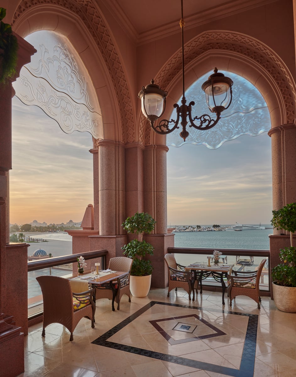 Terrace tables beneath pink stone arches and a wrought-iron lantern, the sea and coast glowing at sunset beyond