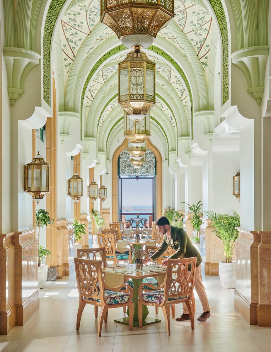 A waiter setting a round table beneath a vaulted pale-green ceiling hung with brass lanterns